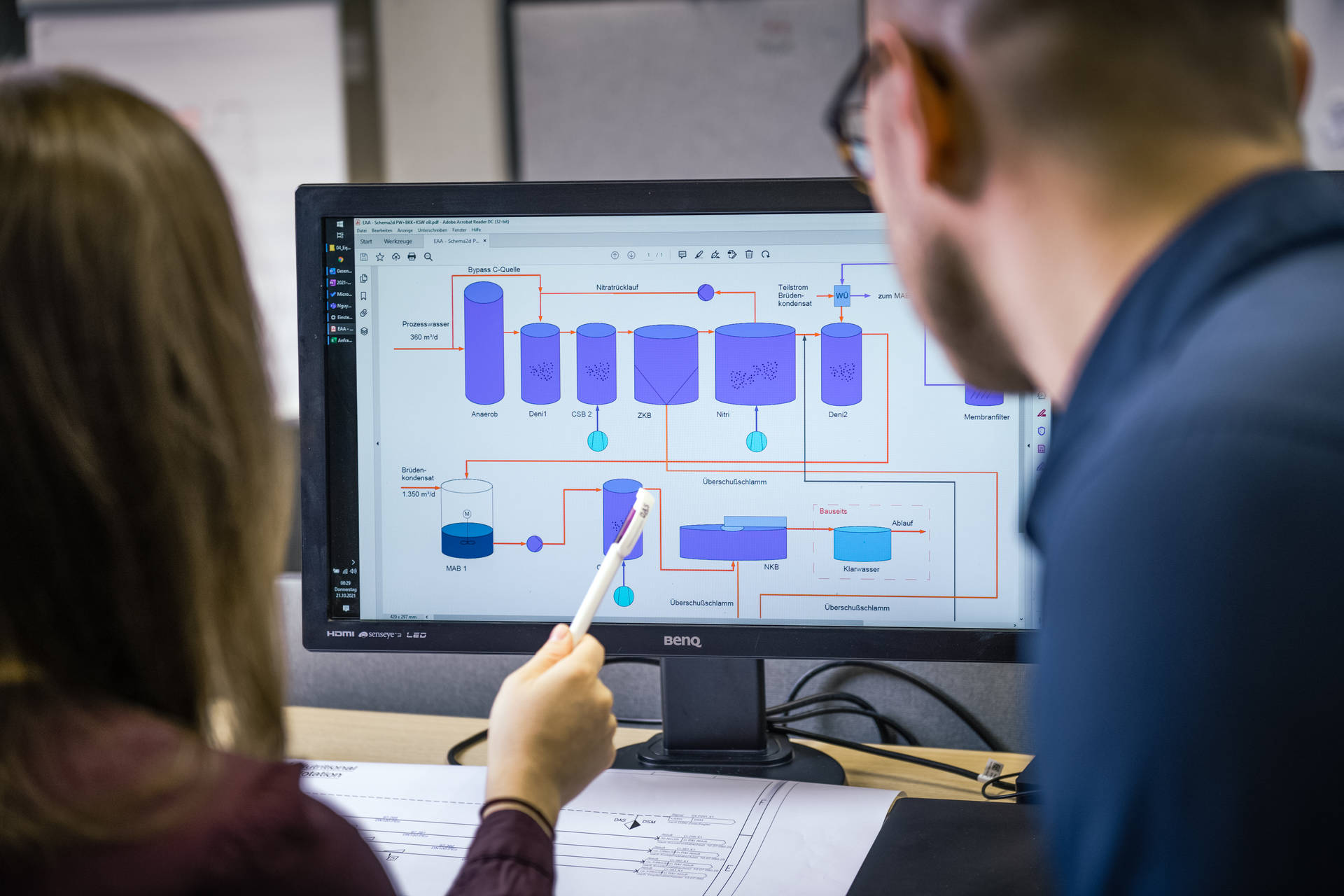 Two people are looking together at a computer screen showing a complex flowchart with various processes and databases, while one person points to part of the diagram with a pen.
