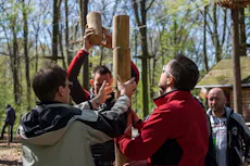 屋外でのチームビルディング活動 森林の中で、数人が木材を使って柱を立てる作業をしている場面。