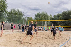 das-volleyballteam A group of people engaging in a lively beach volleyball game on a sandy court, surrounded by trees and a net, under a cloudy sky.