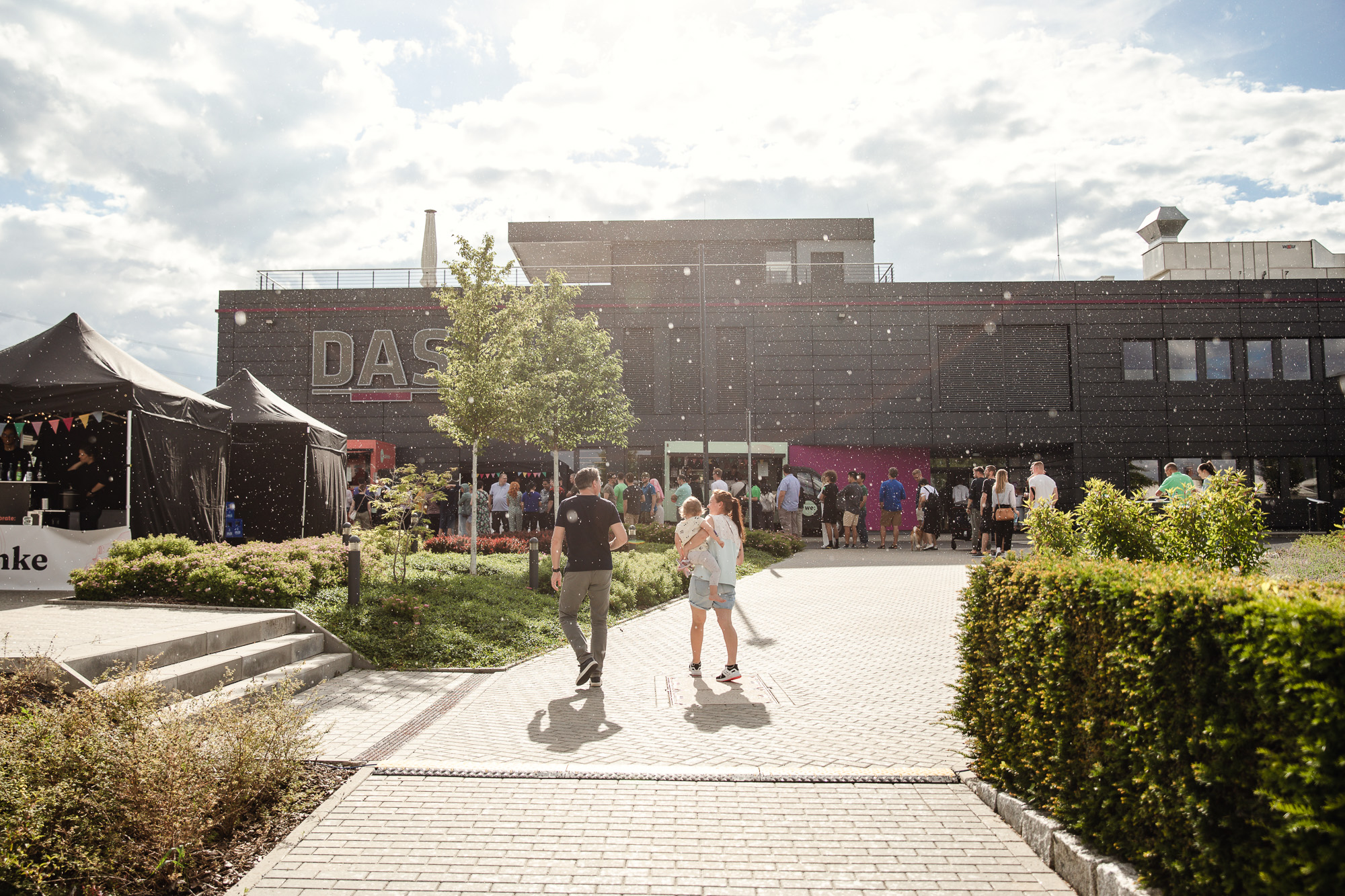 A lively outdoor event at a modern building with people walking on a sunlit pathway and festive tents nearby, surrounded by greenery and a clear sky.