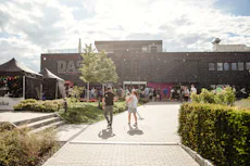 DAS 33.Birthday A lively outdoor event at a modern building with people walking on a sunlit pathway and festive tents nearby, surrounded by greenery and a clear sky.