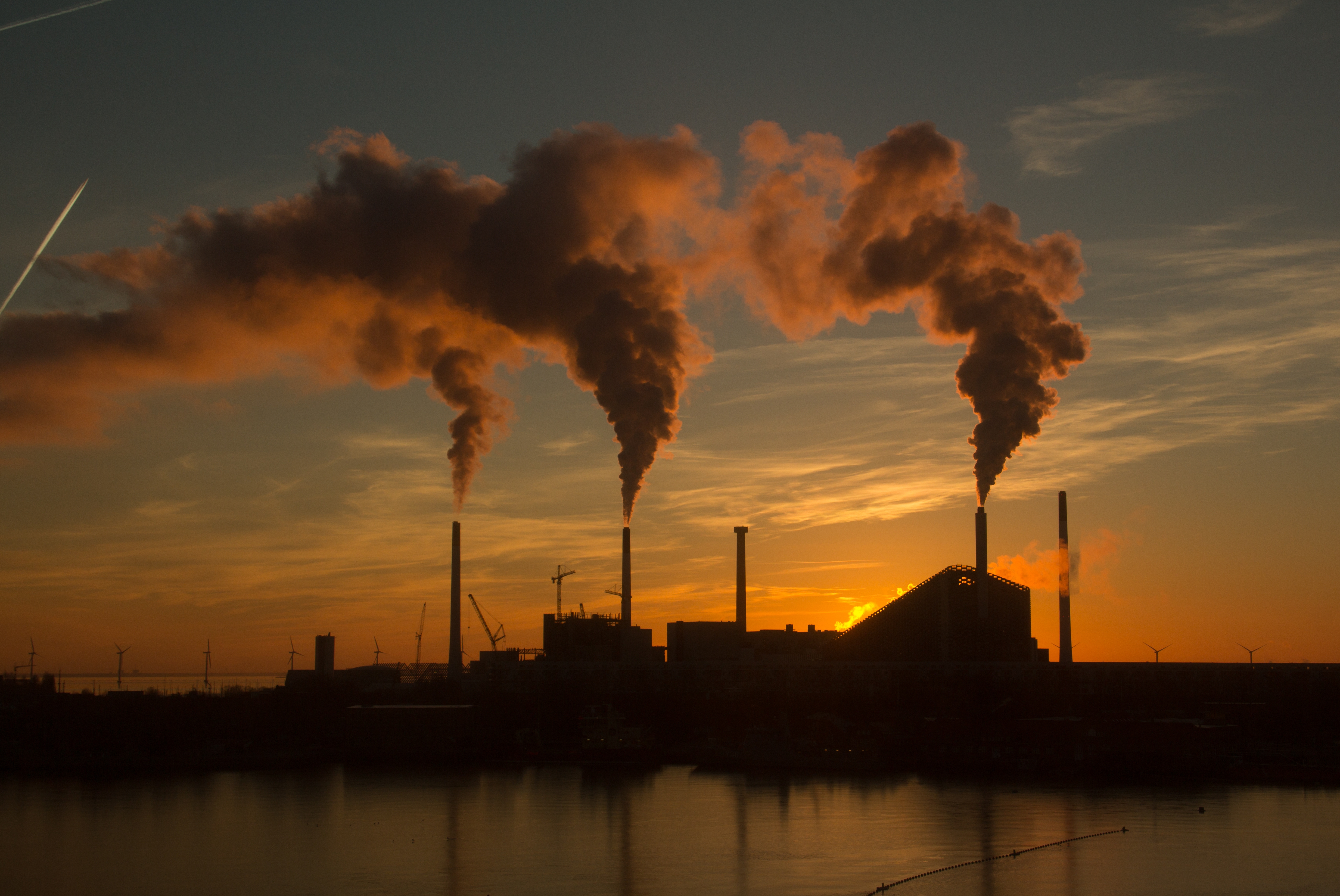 A silhouette of an industrial factory at sunset, emitting thick plumes of smoke from several chimneys into the colorful evening sky, reflecting on a tranquil body of water.