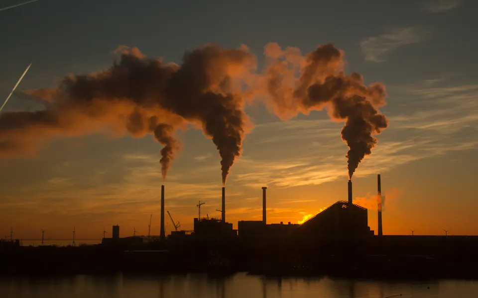 Particulate Matter in the Industry A silhouette of an industrial factory at sunset, emitting thick plumes of smoke from several chimneys into the colorful evening sky, reflecting on a tranquil body of water.