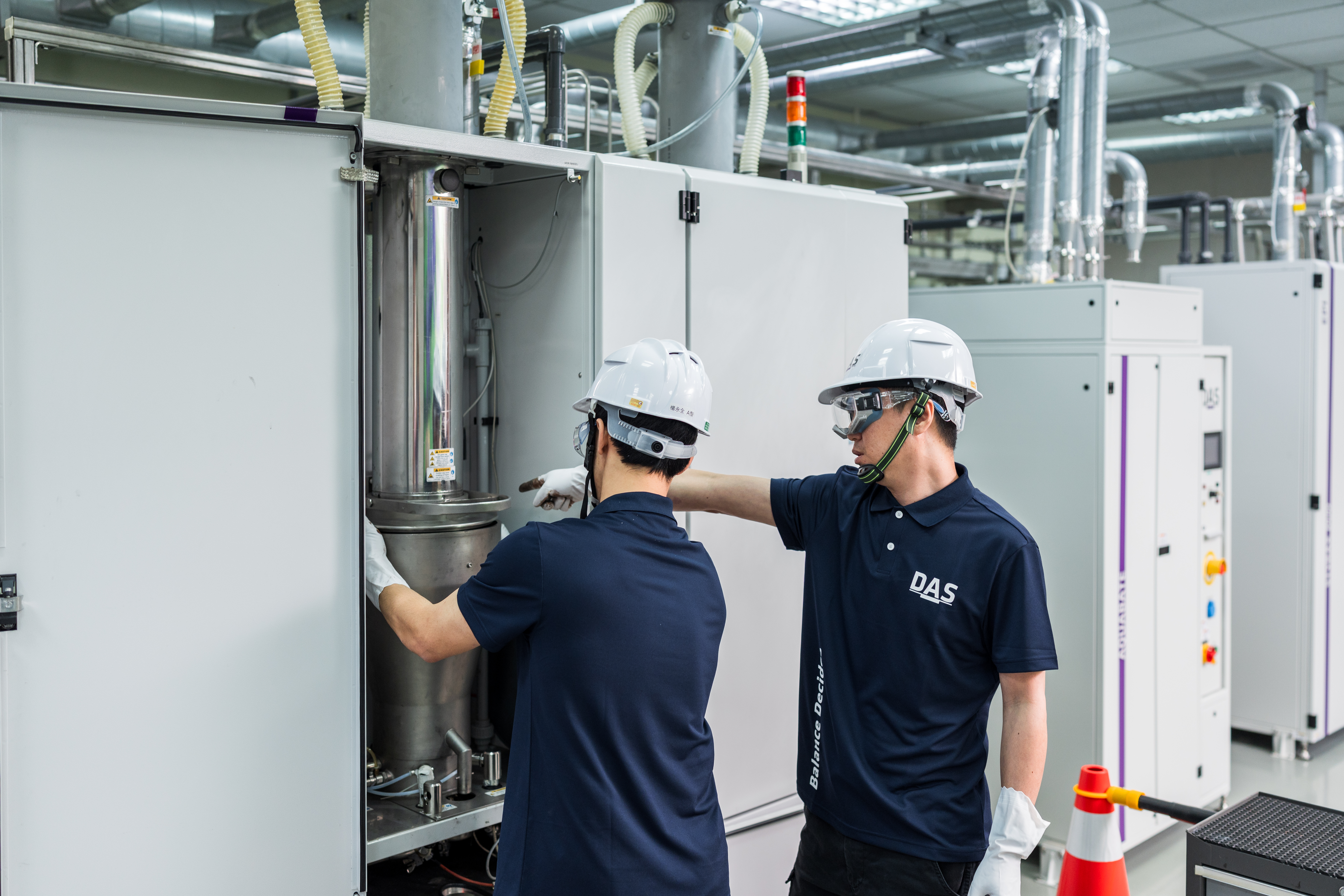 兩名身穿深藍色制服和安全帽的工人在工廠內操作大型機械設備，專注於維修和調整機器的細節。