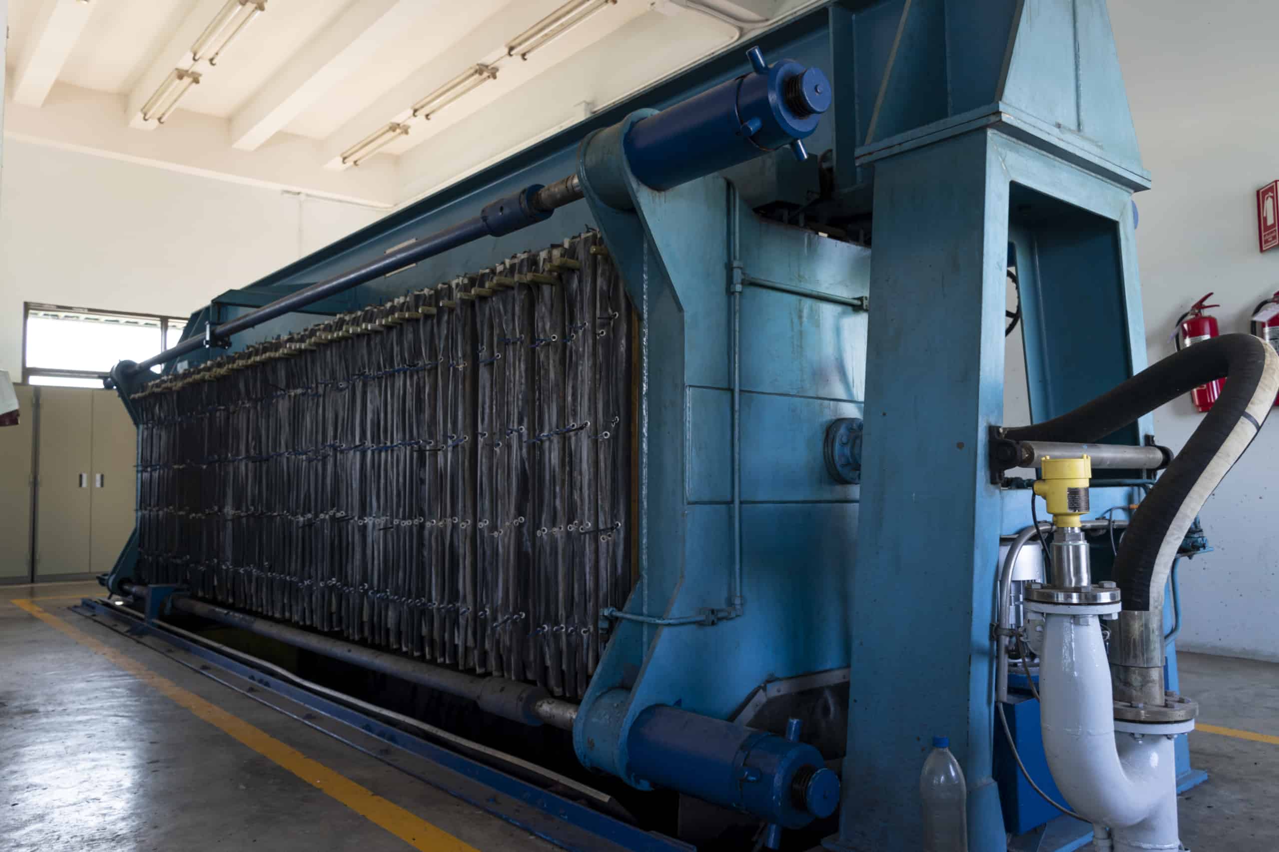 Industrial facility with a large blue filter press standing in a bright room with concrete flooring and neon ceiling lights, surrounded by pipes and machine components.