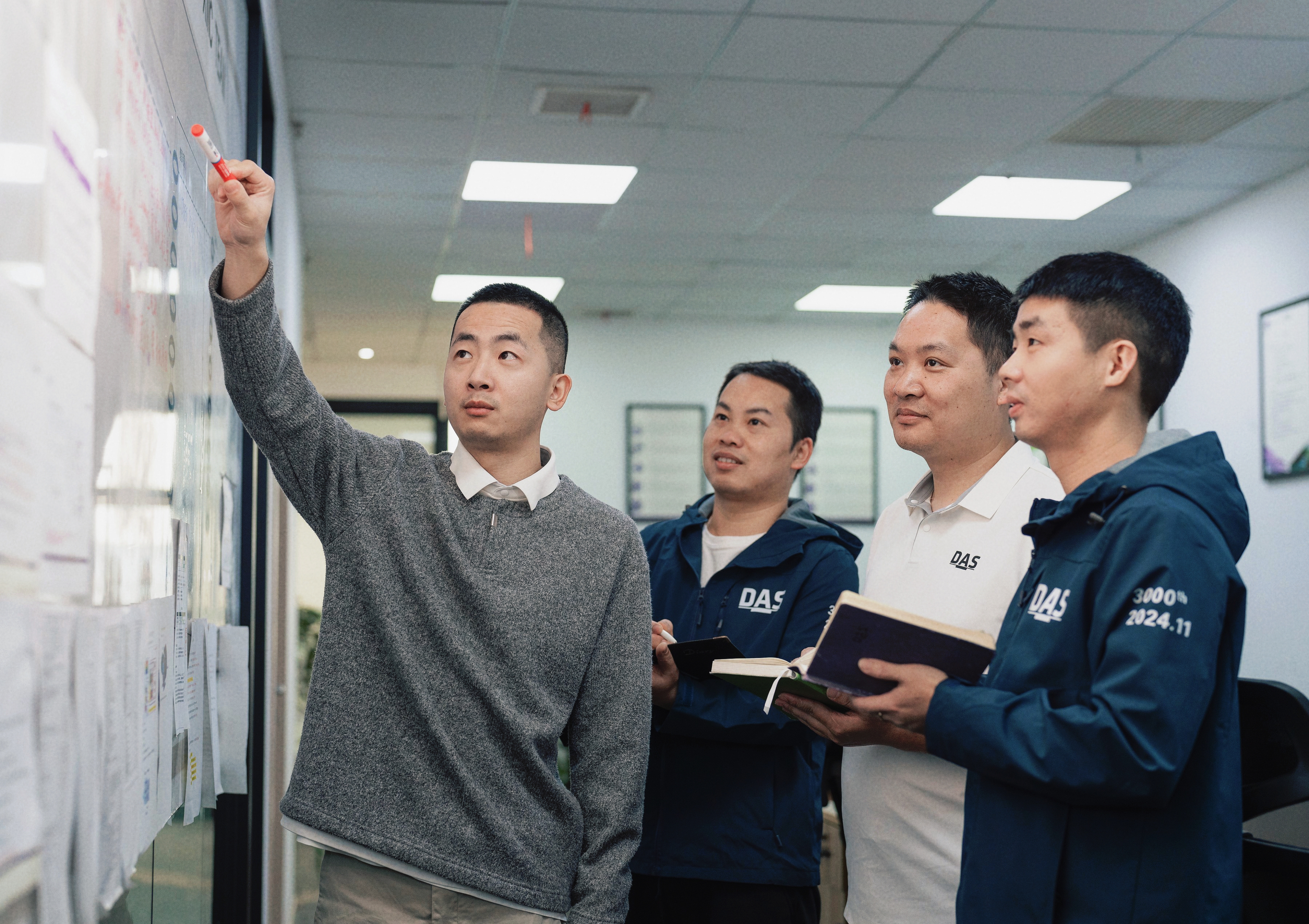 Four individuals are in an office, with one pointing at a whiteboard covered in charts and papers, while the others observe attentively, all dressed in casual business attire.