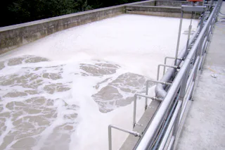 Reference Paper Industry Aerial view of an industrial wastewater treatment facility with a rectangular layout, showcasing large tanks filled with sediment in varying shades of brown, surrounded by pipes and machinery, set against a backdrop of trees.