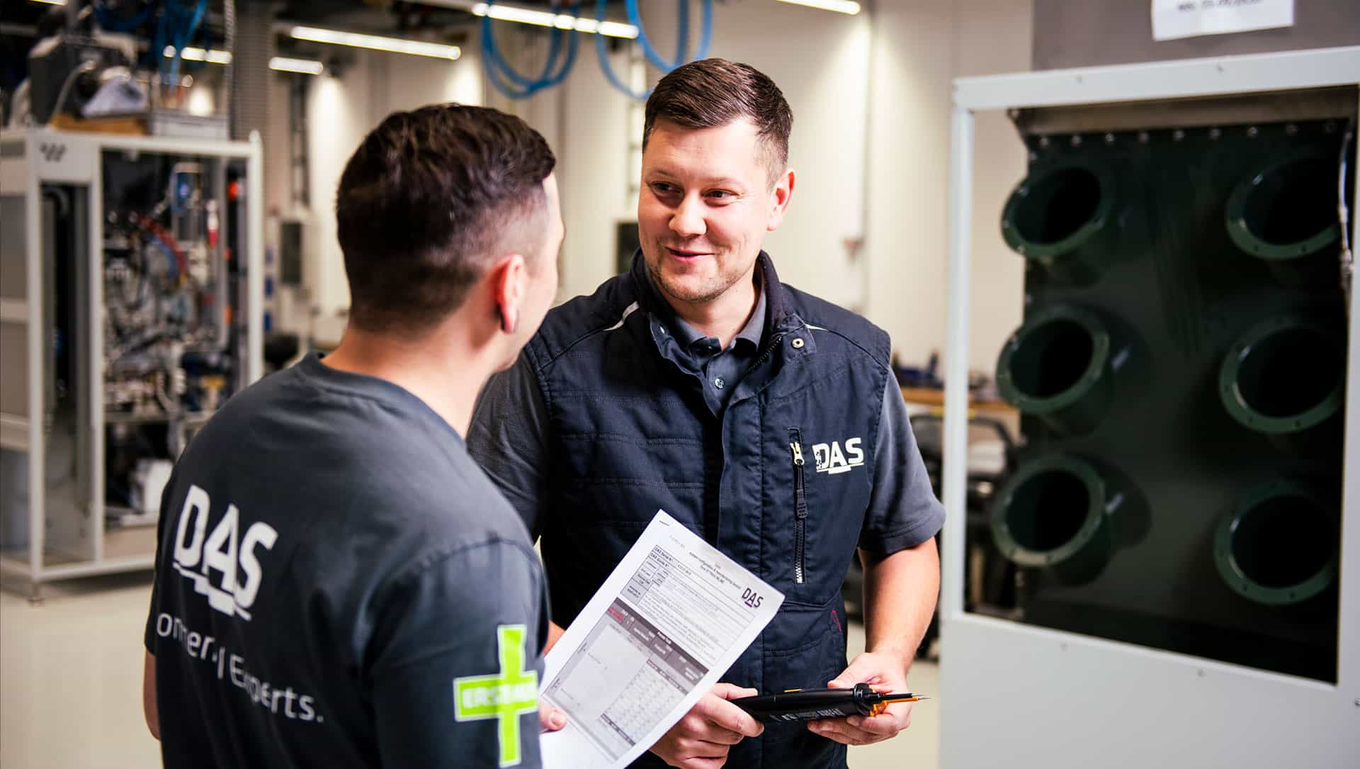Two men are talking in a modern industrial workshop, one holding a form and the other carrying a tablet, surrounded by machines and technical equipment.