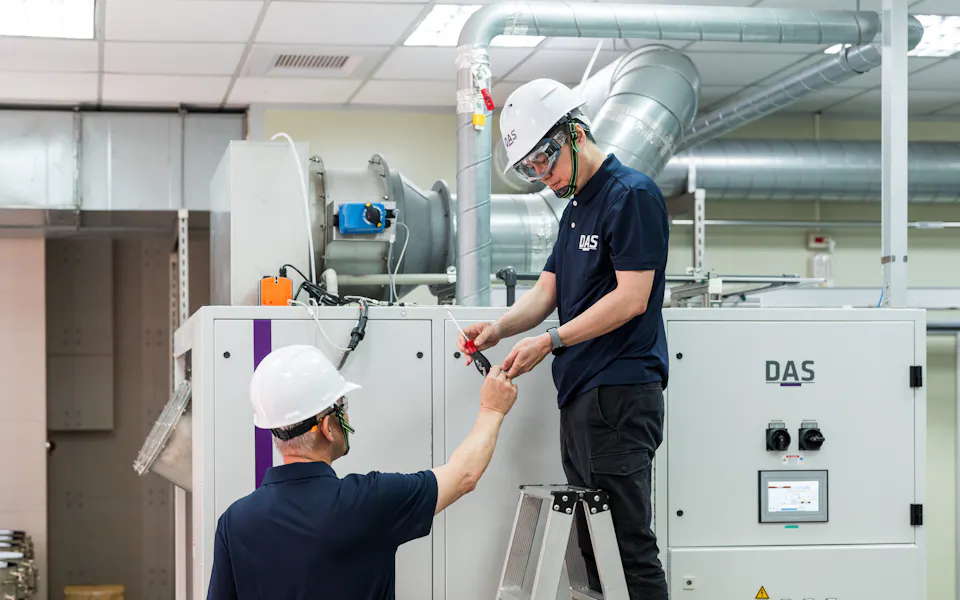 機台檢測-1 兩位穿著工作服和安全帽的工人在工廠內操作機器設備,一人站在梯子上進行調整,另一人則在地面協助。