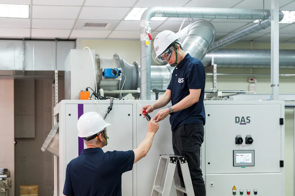 兩位穿著工作服和安全帽的工人在工廠內操作機器設備，一人站在梯子上進行調整，另一人則在地面協助。