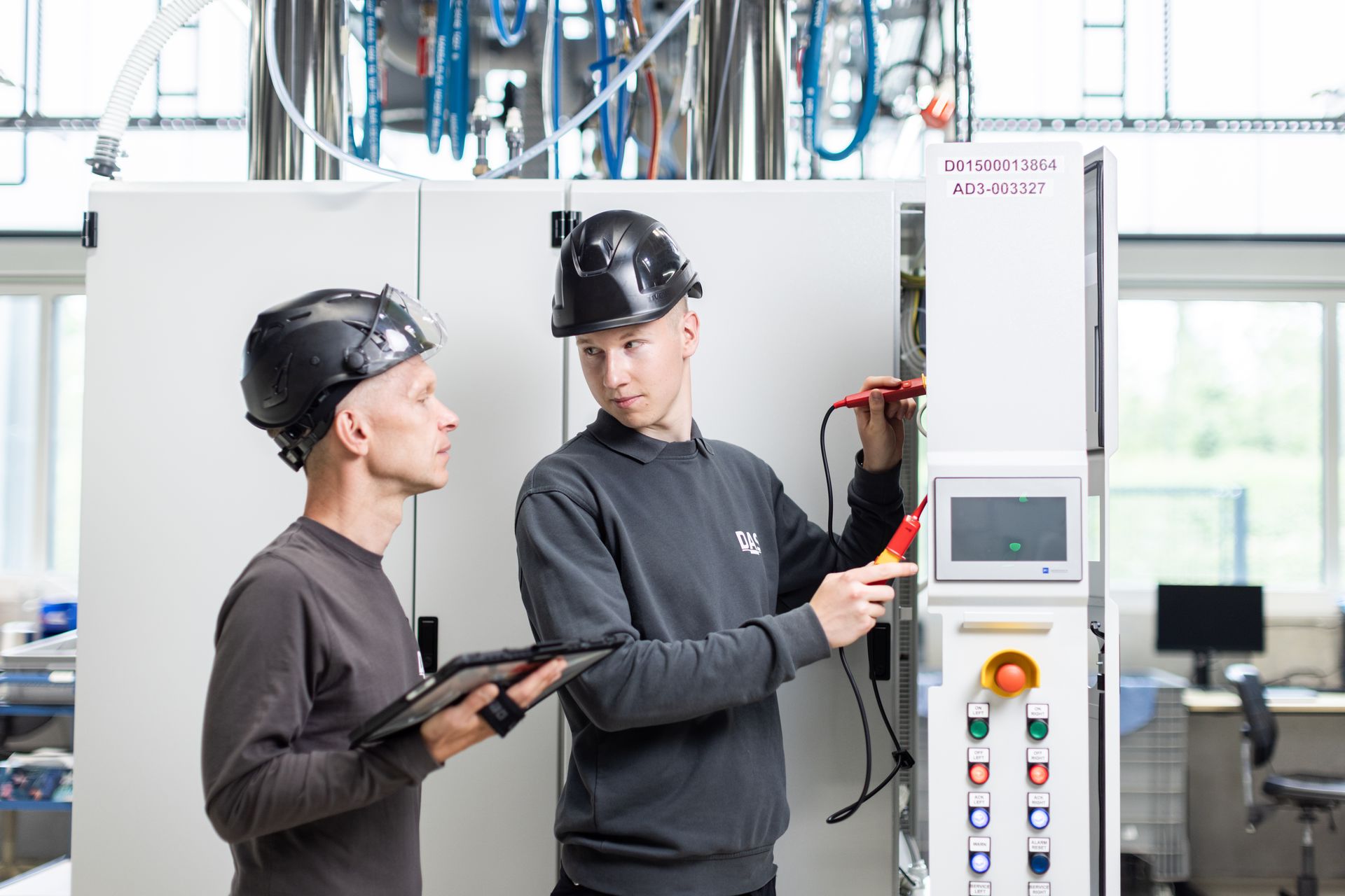 Two technicians wearing safety helmets work on an industrial control panel inside a modern manufacturing facility, with one holding a tablet and the other using testing equipment.