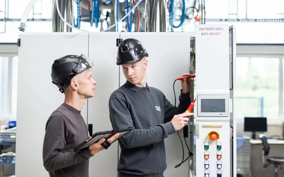 Service team working on treatment system Two technicians wearing safety helmets work on an industrial control panel inside a modern manufacturing facility, with one holding a tablet and the other using testing equipment.