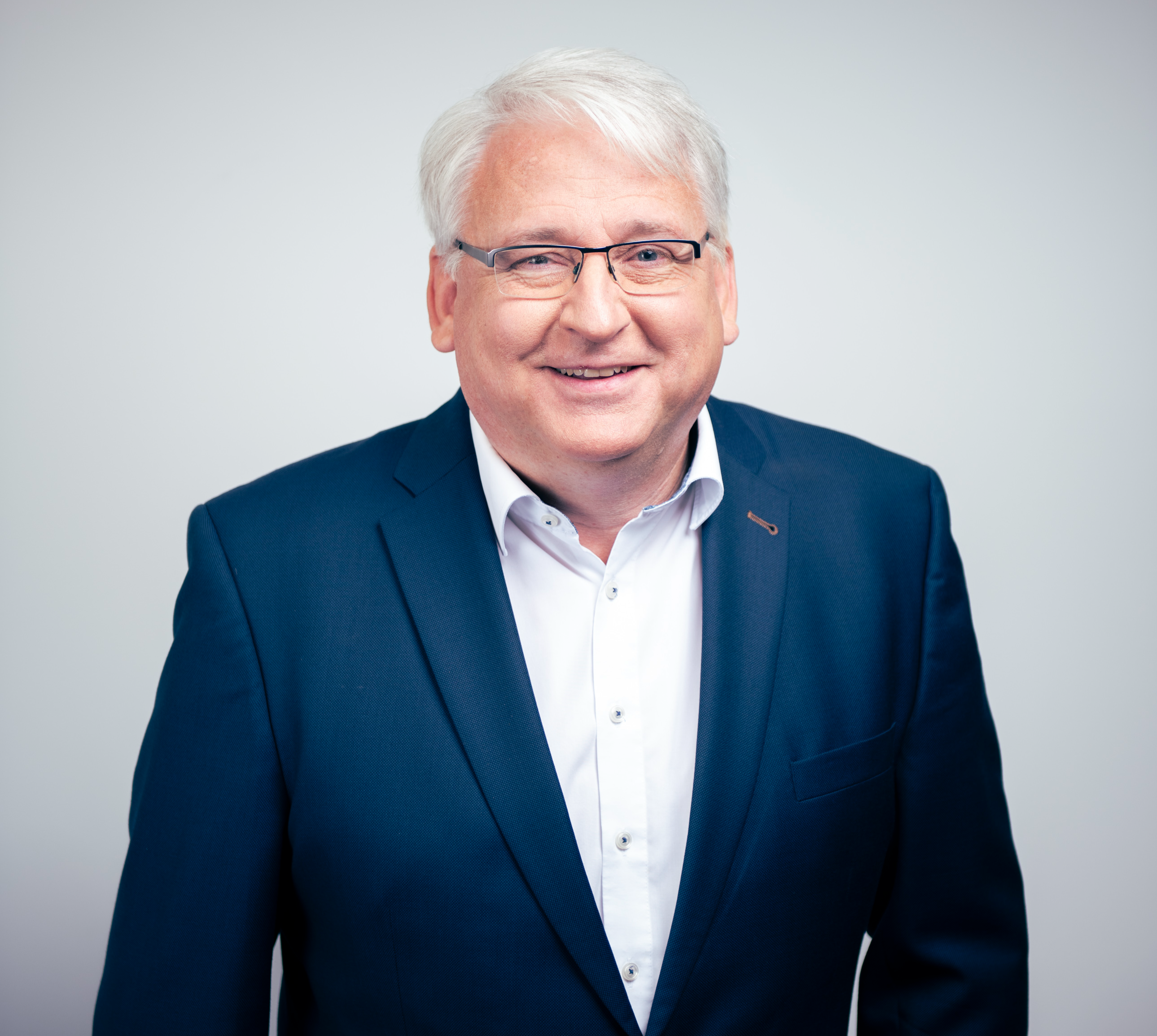 An older man with white hair is smiling, wearing a navy blue suit over a white dress shirt, against a plain, light-colored background.