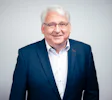 Dr. Guy Davies, Chief Business Development Officer An older man with white hair is smiling, wearing a navy blue suit over a white dress shirt, against a plain, light-colored background.