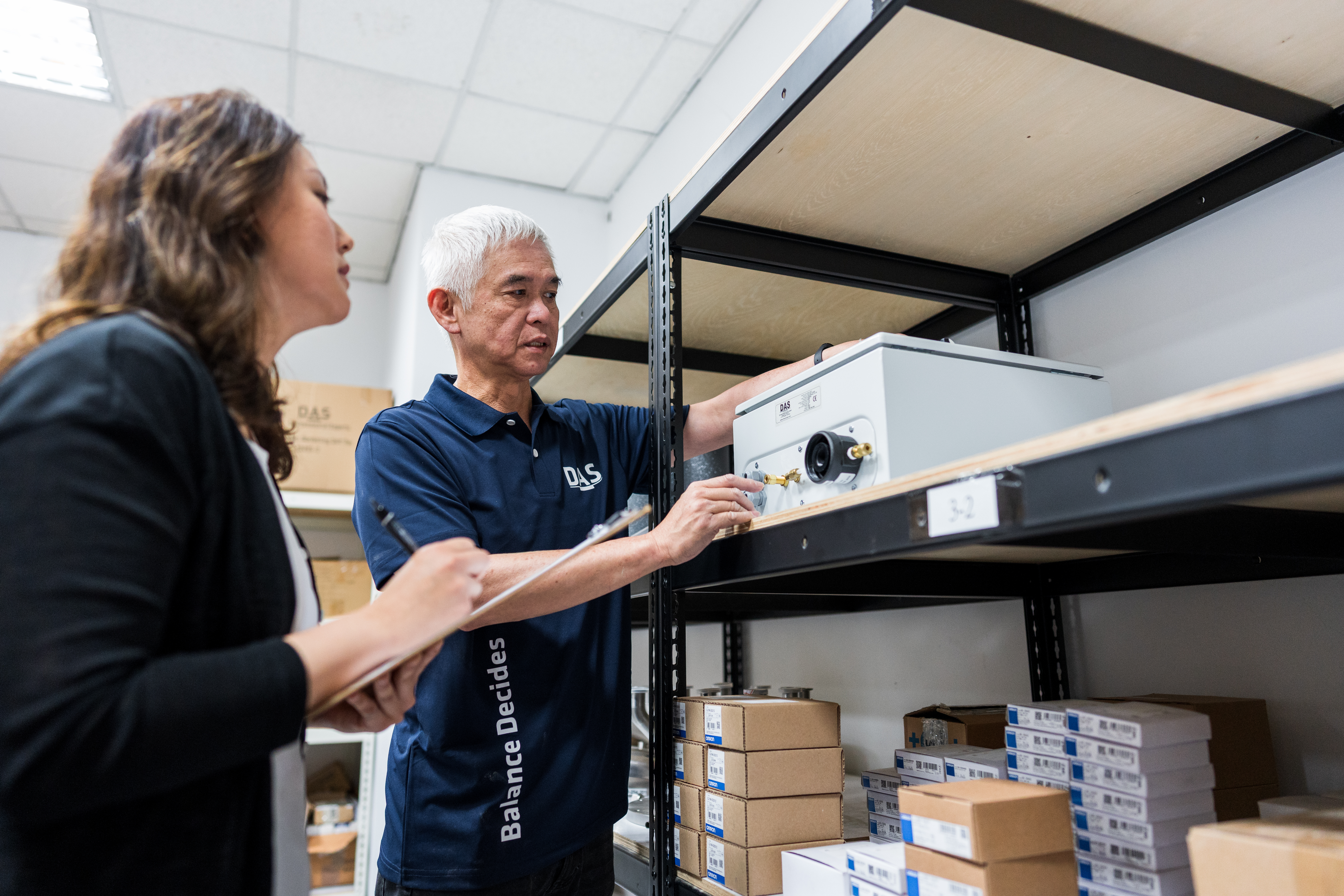 兩位同事站在倉庫裡，正在討論放在架子上的設備，一位寫筆記，周圍有幾個裝滿物品的紙箱。