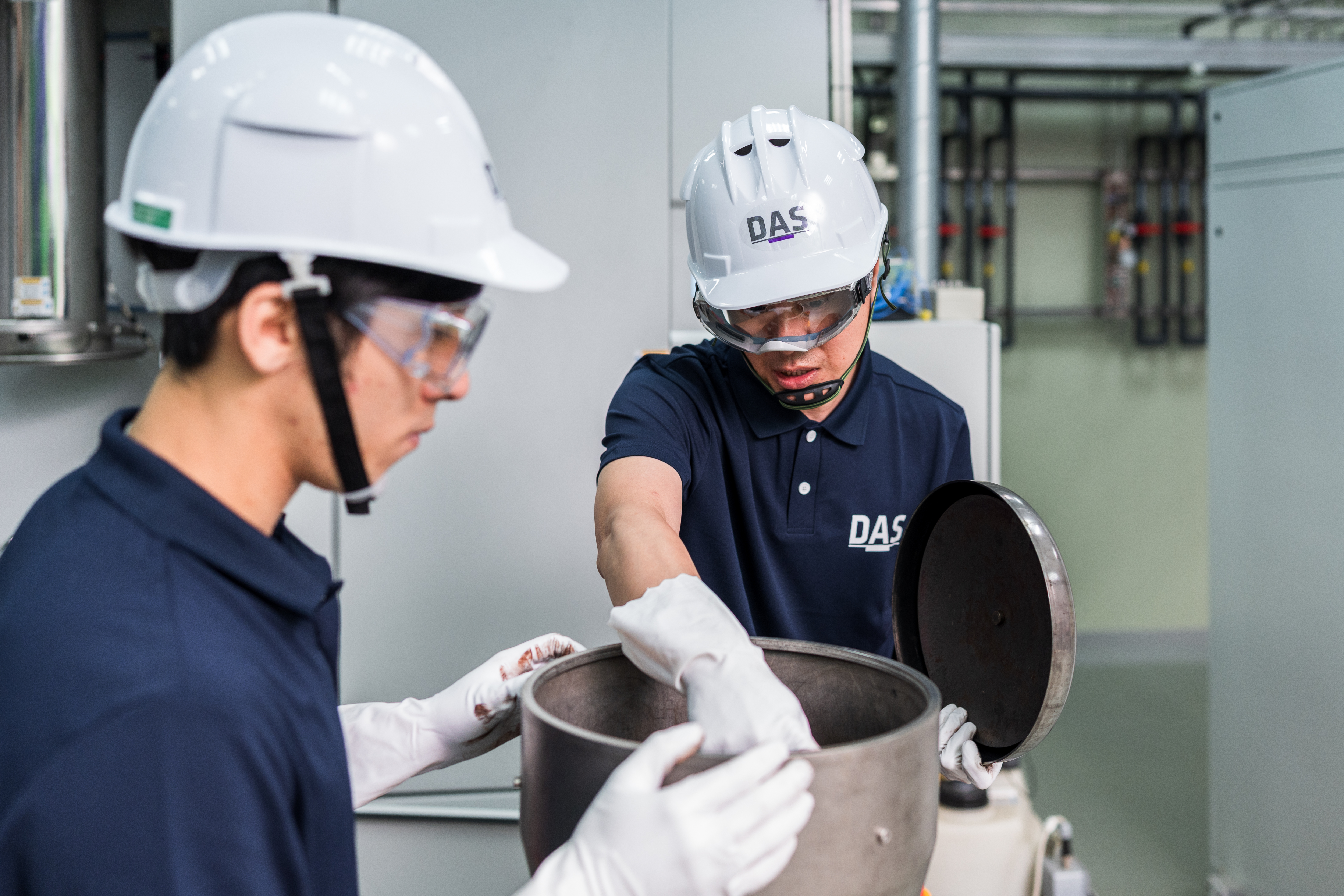 兩名身穿深藍色制服和白色安全帽的工人正在工廠內進行設備檢查，其中一位工人正在專注地檢視金屬容器內部。