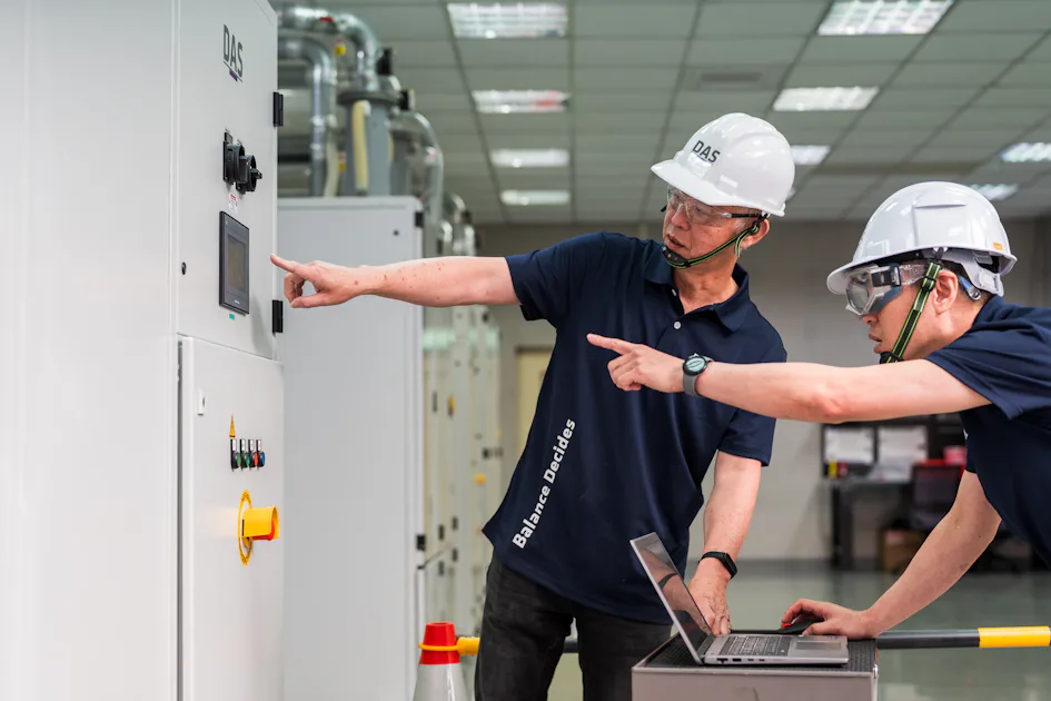 數據討論 兩名戴著安全帽的技術人員站在工廠車間內查看大型白色控制面板,其中一人正在操作筆記型電腦。