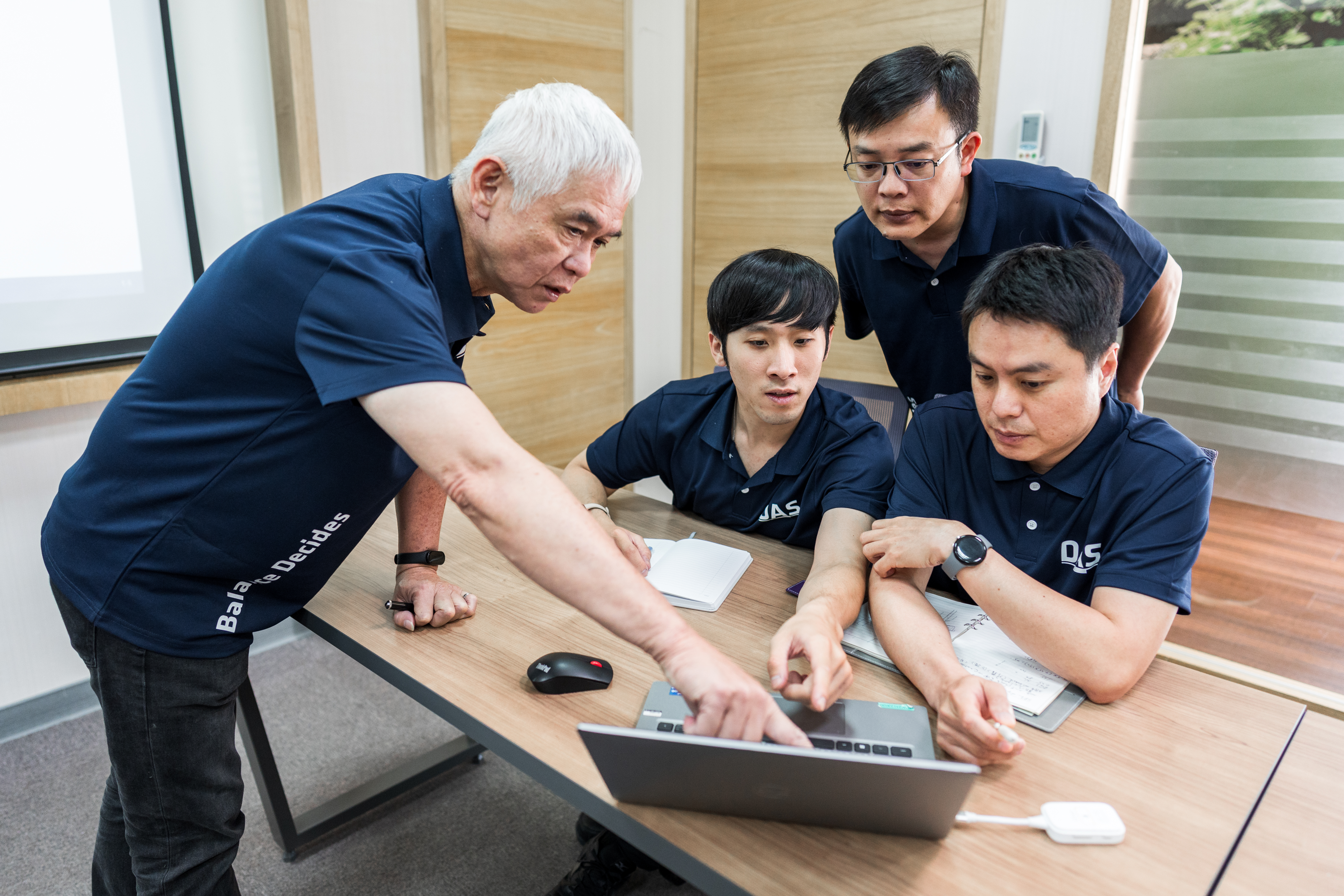 一位年長男性正在向坐在桌子前的三位同事展示筆記型電腦螢幕中的內容，辦公室環境下進行互動與討論。