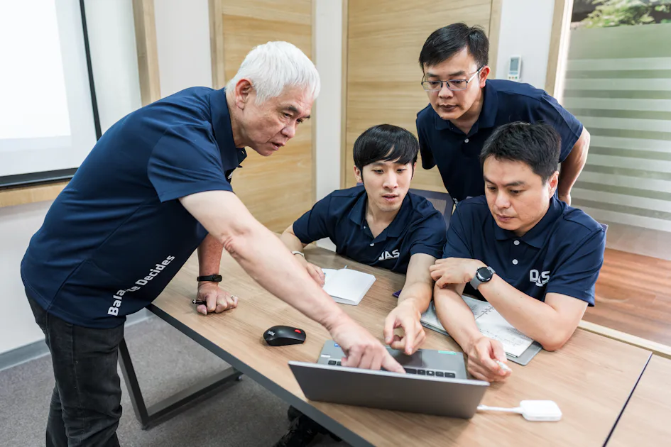 分析討論 一位年長男性正在向坐在桌子前的三位同事展示筆記型電腦螢幕中的內容,辦公室環境下進行互動與討論。