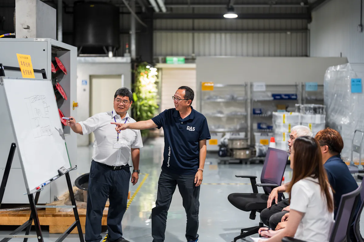 工廠開會 在一間工廠的會議室裡,兩位穿著休閒服的男子正在白板上進行演示,白板上有草圖,幾位坐著的聽眾在關注他們,背景包含架子和一些工業設備。