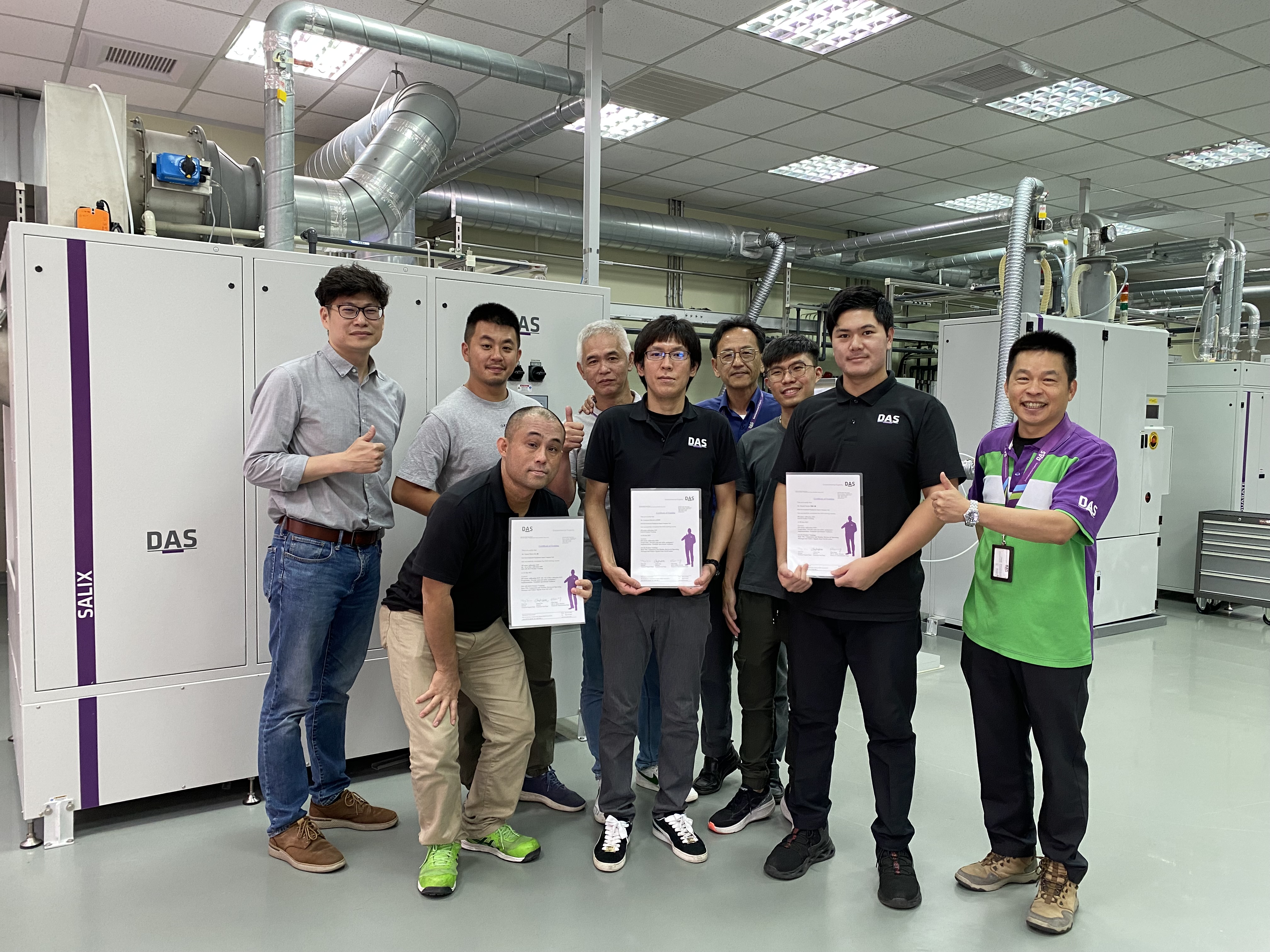 A group of people stand together in a factory setting, with some holding certificates, in front of large industrial machinery, indicating a professional or award ceremony.