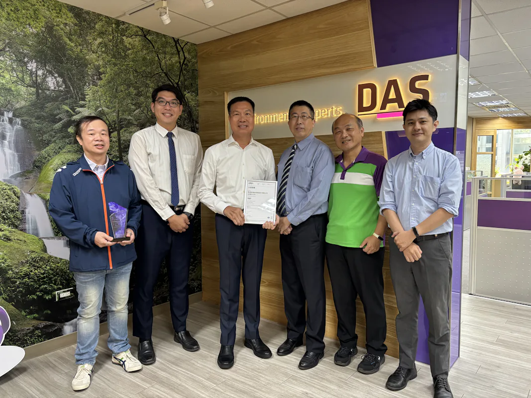 ISO 授證儀式 一群身穿商務休閒服裝的男性在辦公室的接待區合影,背景是帶有公司標誌的紫色牆面和自然風景壁紙。