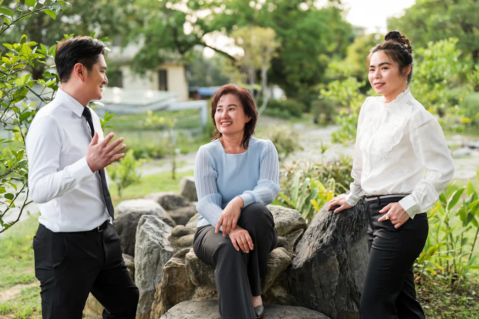 將軍村外景 三位穿著正式商務服裝的人在綠意盎然的戶外環境中微笑交流,其中一位坐在石頭上,其餘兩位站著。