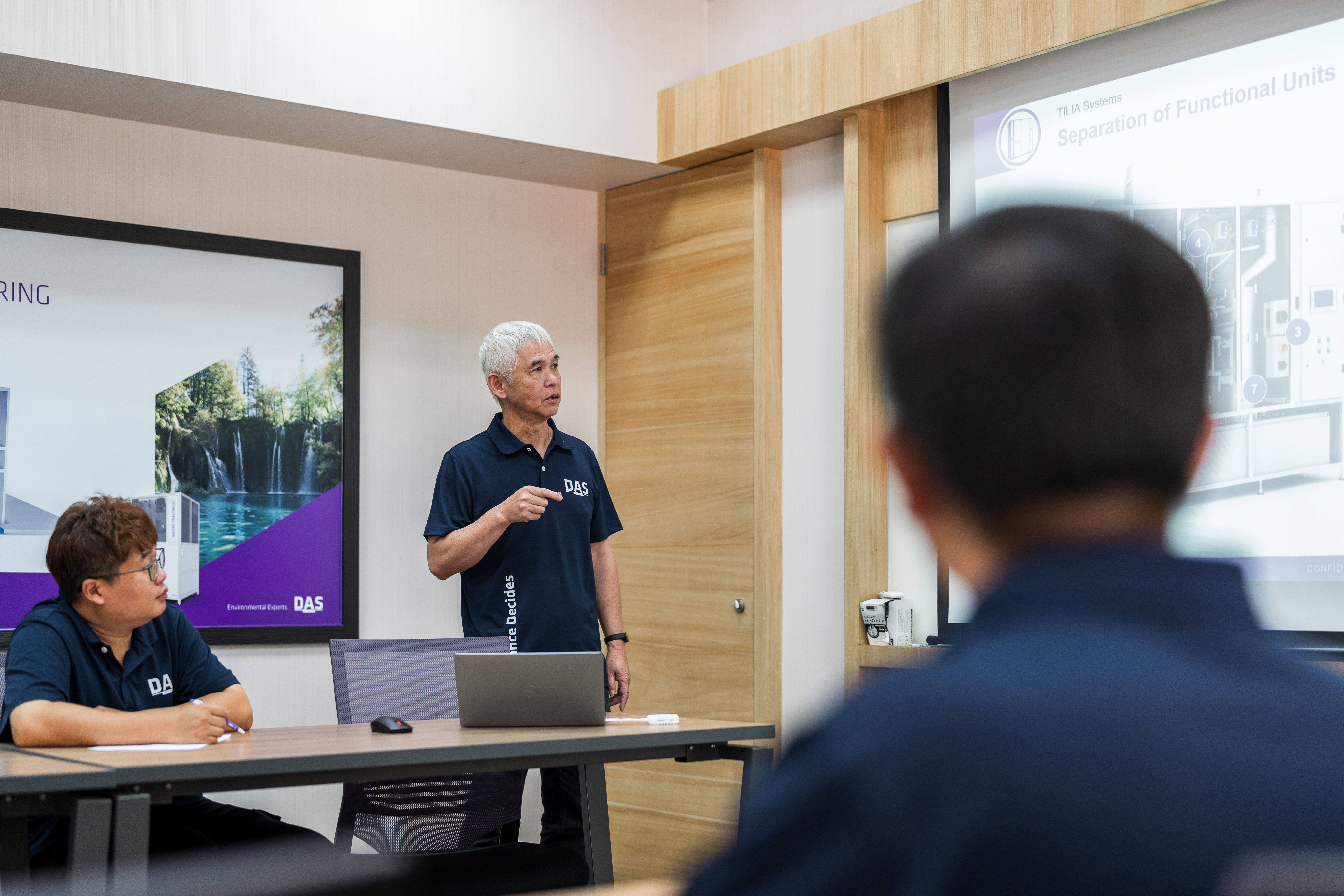 一名穿著藍色襯衫的男子在會議室裡進行演示，背景有顯示技術圖表的大螢幕，幾位參加者在桌子周圍坐著專心聆聽。