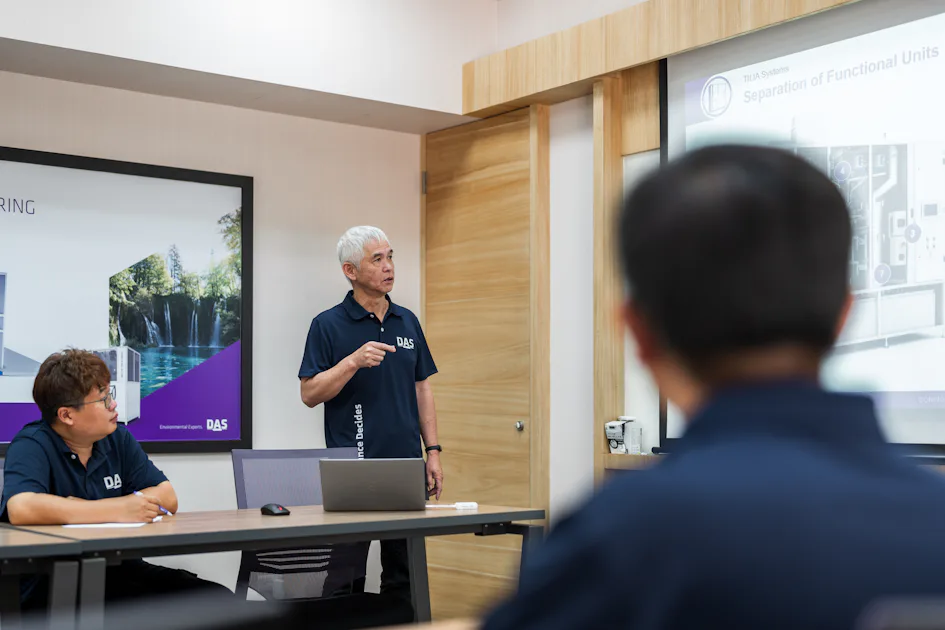 ISC training 一名穿著藍色襯衫的男子在會議室裡進行演示,背景有顯示技術圖表的大螢幕,幾位參加者在桌子周圍坐著專心聆聽。