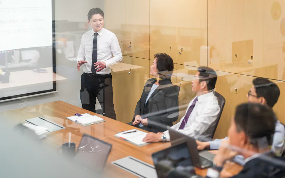 討論 一名穿著白襯衫的商務人士在會議室內向坐在桌邊的其他商務人士進行演示,桌上擺放著筆記型電腦和文件,展示出專業的工作環境。