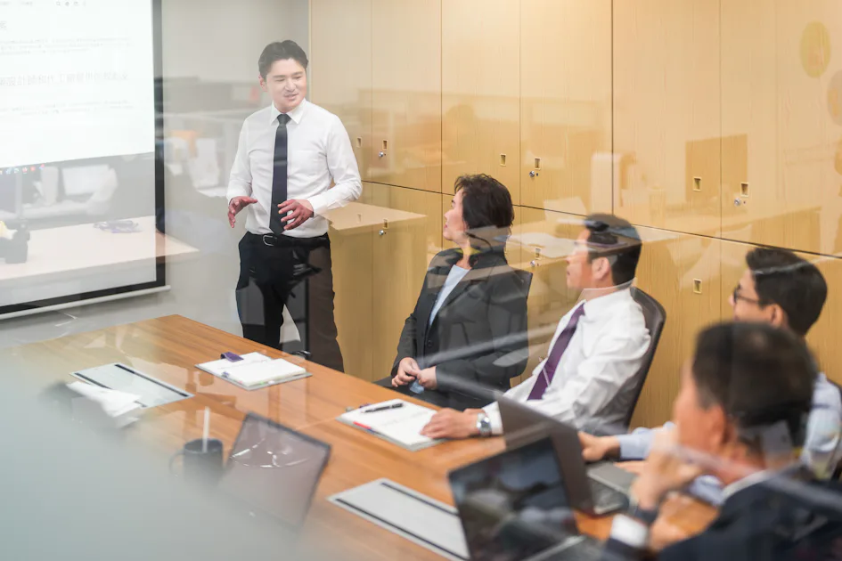 討論 一名穿著白襯衫的商務人士在會議室內向坐在桌邊的其他商務人士進行演示,桌上擺放著筆記型電腦和文件,展示出專業的工作環境。