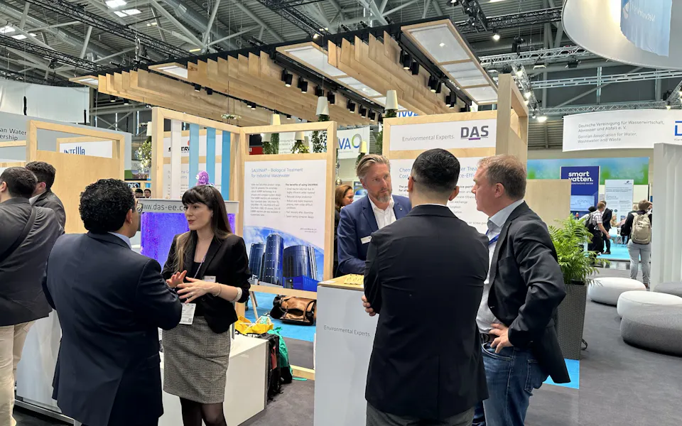 Impressions IFAT A busy trade show booth at a convention center features four men and one woman engaged in animated discussions, surrounded by wooden structures and bright informational displays with hanging lights, while other exhibition stalls are visible in the background.