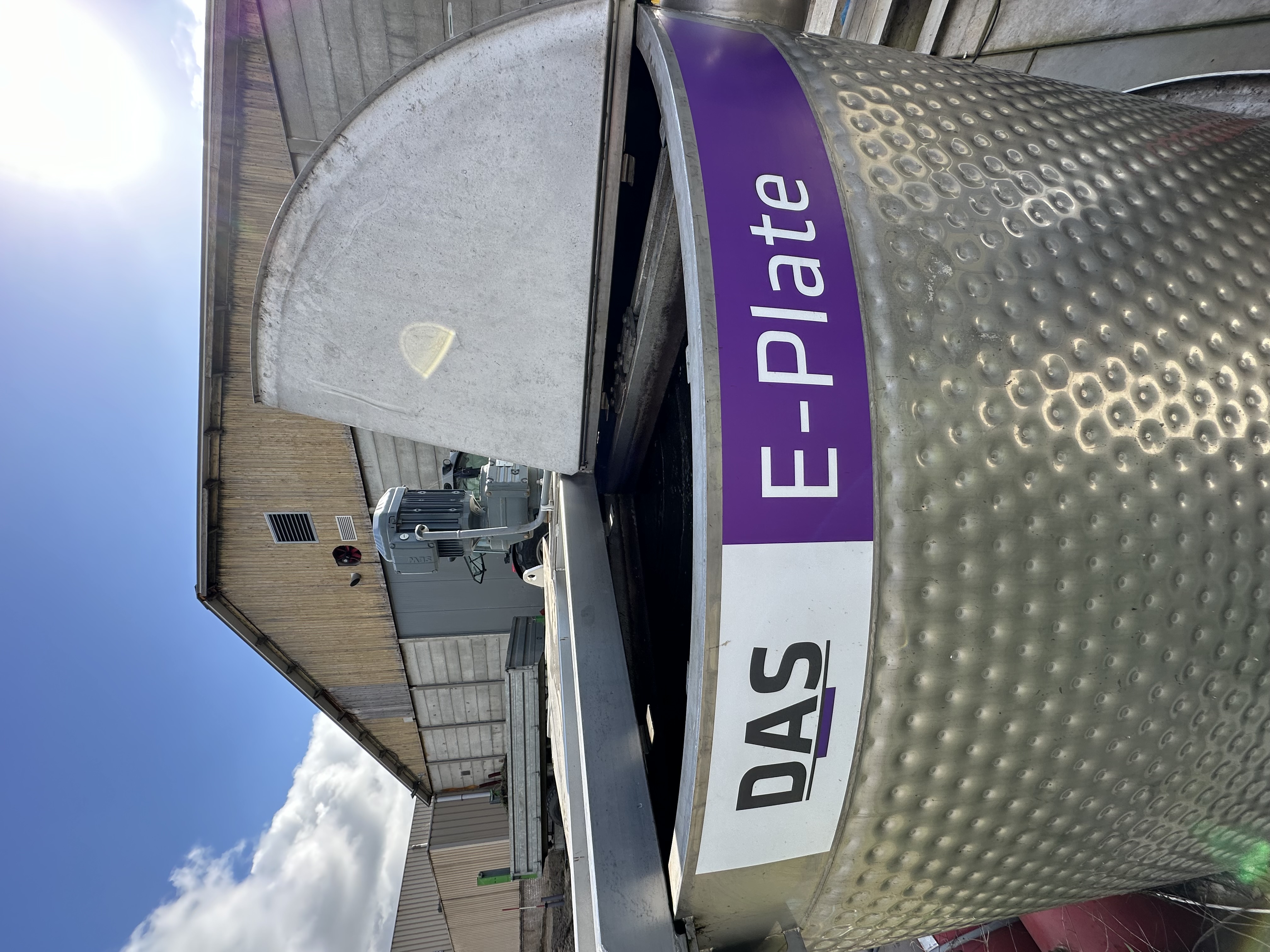 A stainless-steel E-Plate system with a partially open lid is situated outdoors, with a background of industrial buildings and a clear blue sky.