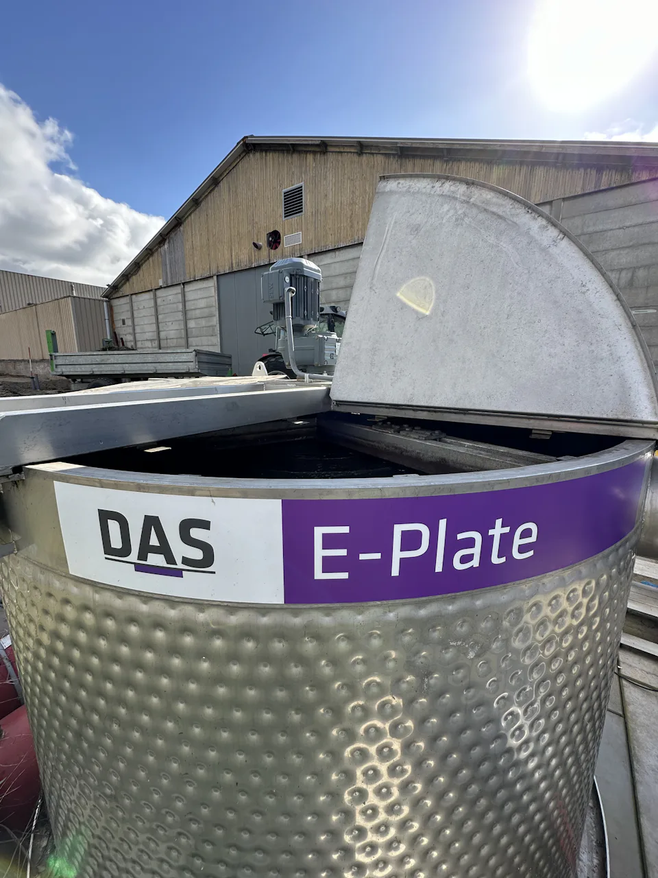 A stainless-steel E-Plate system with a partially open lid is situated outdoors, with a background of industrial buildings and a clear blue sky.
