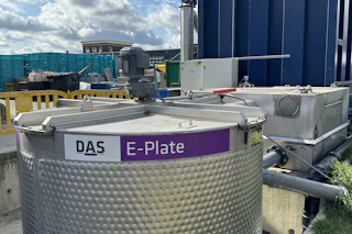 E-PLATE bei Evers Specials B.V. Ein großer Edelstahlbehälter mit der Aufschrift „E-Plate“ steht im Freien unter einem teilweise bewölkten Himmel, angebunden an ein Rohrleitungssystem und weitere Anlagen.