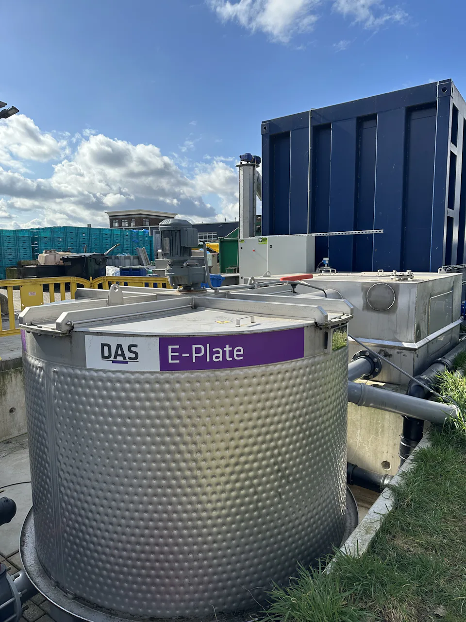 E-PLATE at Evers Specials B.V. A large stainless steel container labelled ‘E-Plate’ stands outdoors under a partly cloudy sky, connected to a pipe system and other equipment.