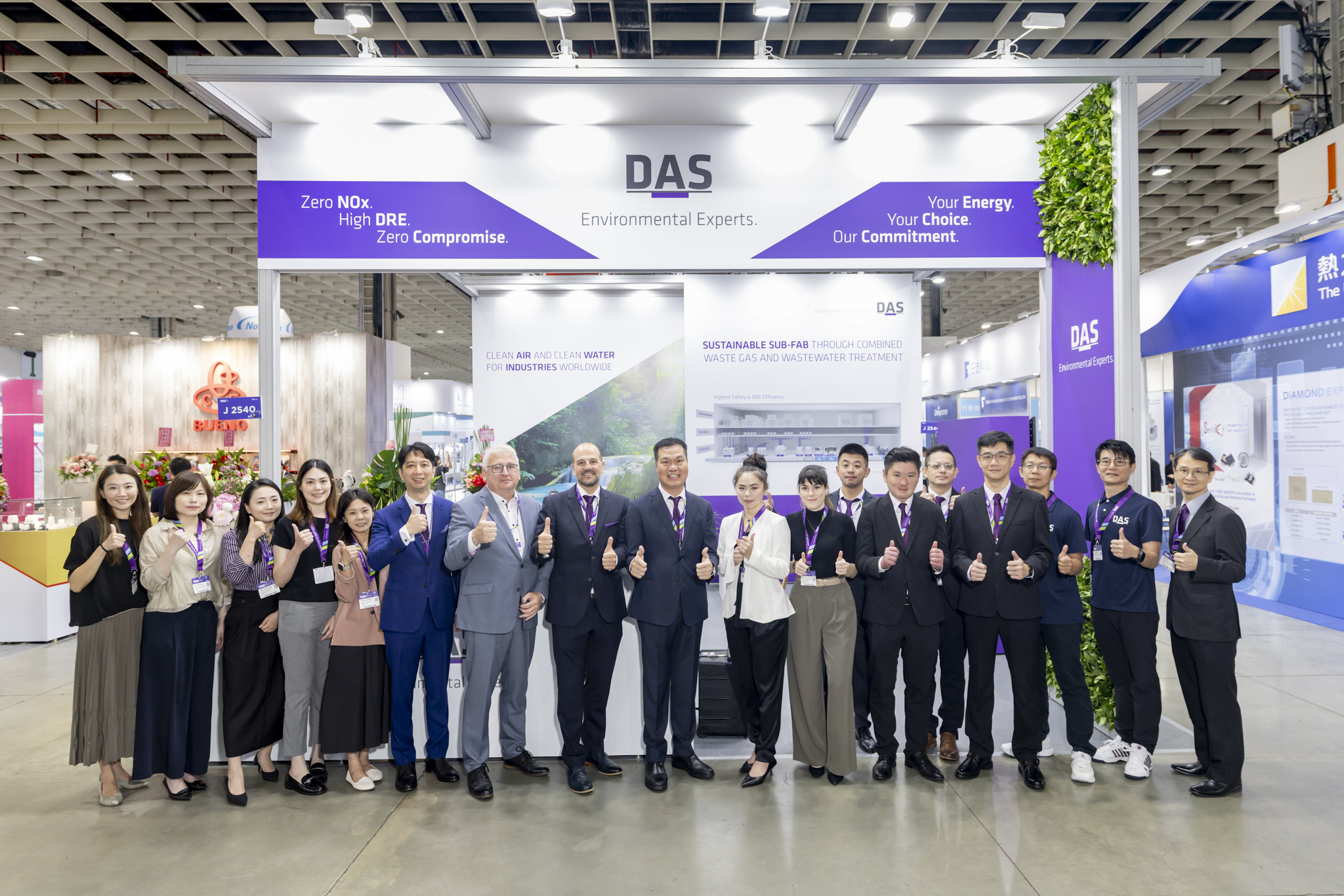 A group of people stand in front of a trade show booth labeled "DAS Environmental Experts," all giving thumbs up, with banners promoting environmental solutions in the background.