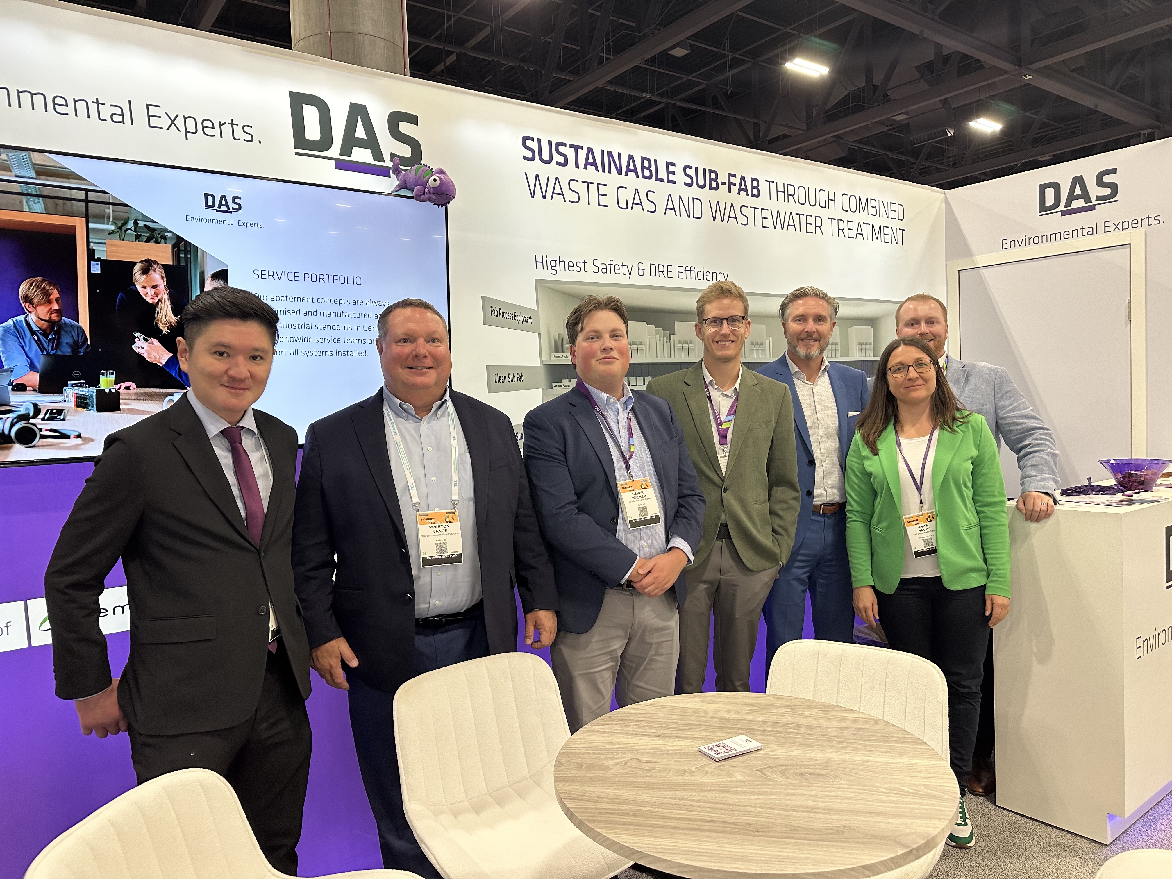 A group of people is standing in a business exhibit booth featuring a display about "Sustainable Sub-Fab Through Combined Waste Gas and Wastewater Treatment" by DAS Environmental Experts, with a modern design, informational banners, and a round table in the foreground.