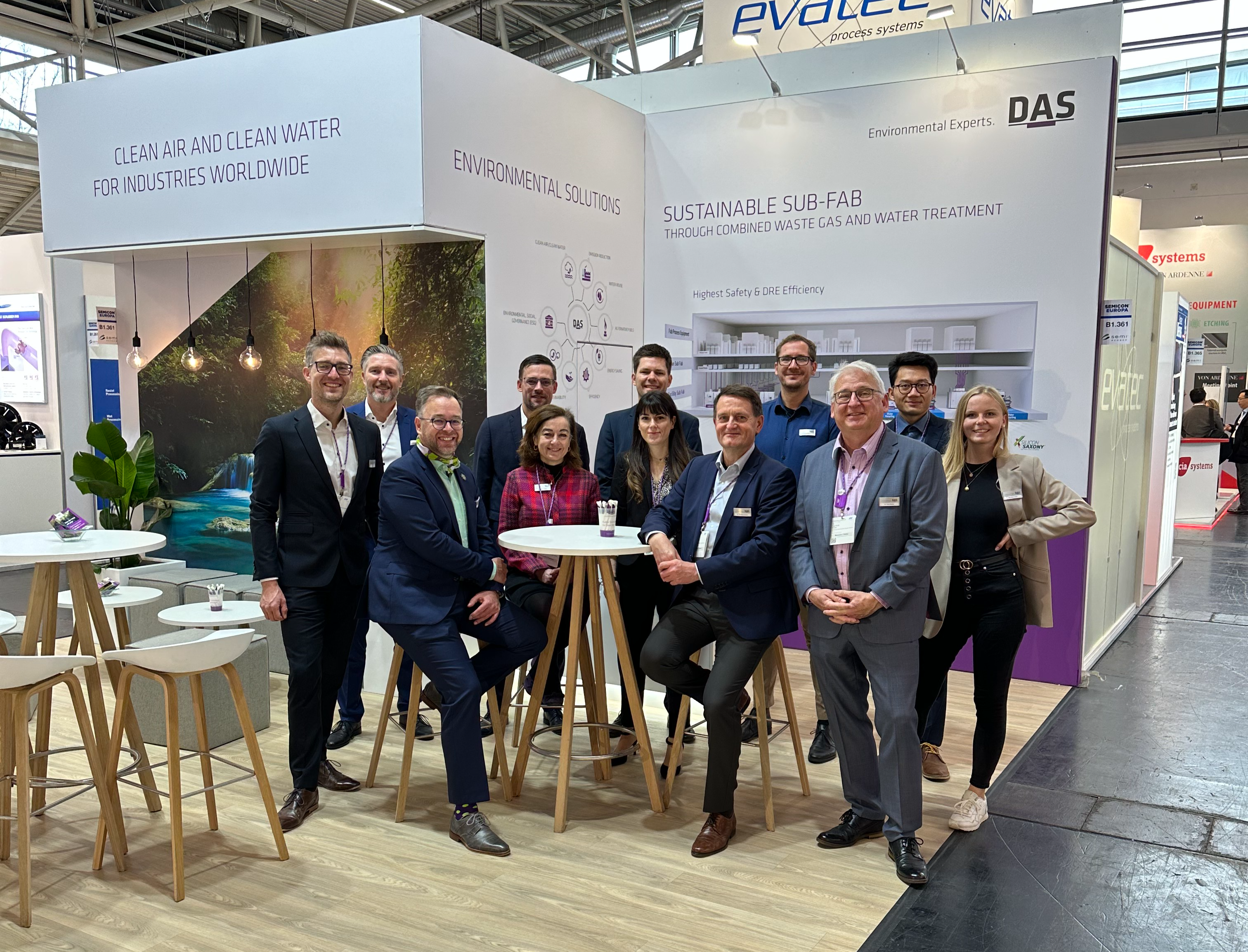 A group of people in professional attire pose together at a trade show booth featuring modern design elements, with displays and branding from DAS visible in the background.