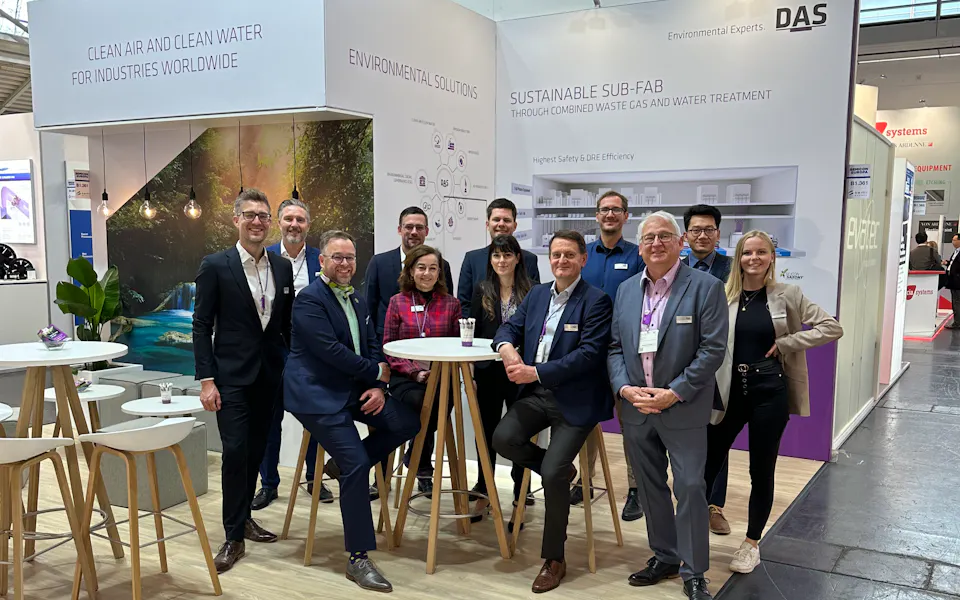 Impressions SEMICON Europa A group of people in professional attire pose together at a trade show booth featuring modern design elements, with displays and branding from DAS visible in the background.
