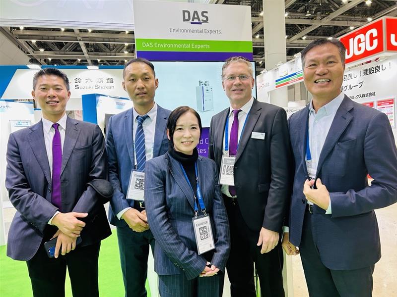 Five people are standing together inside a convention center, wearing business attire, with a banner for DAS Environmental Experts visible in the background.
