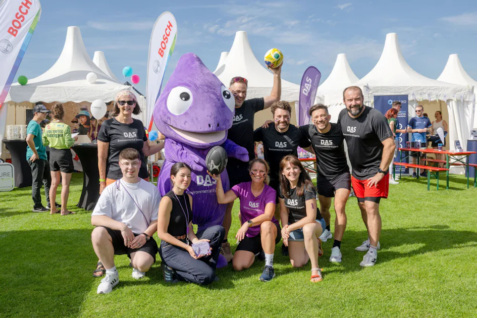 1st Silicon Saxony Sports Day A group of people wearing matching black shirts pose for a cheerful photo with a large purple mascot, set against a backdrop of white tents and flags under a clear blue sky at an outdoor event.