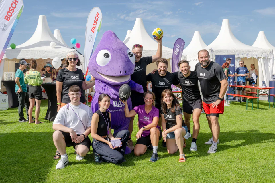 1st Silicon Saxony Sports Day A group of people wearing matching black shirts pose for a cheerful photo with a large purple mascot, set against a backdrop of white tents and flags under a clear blue sky at an outdoor event.