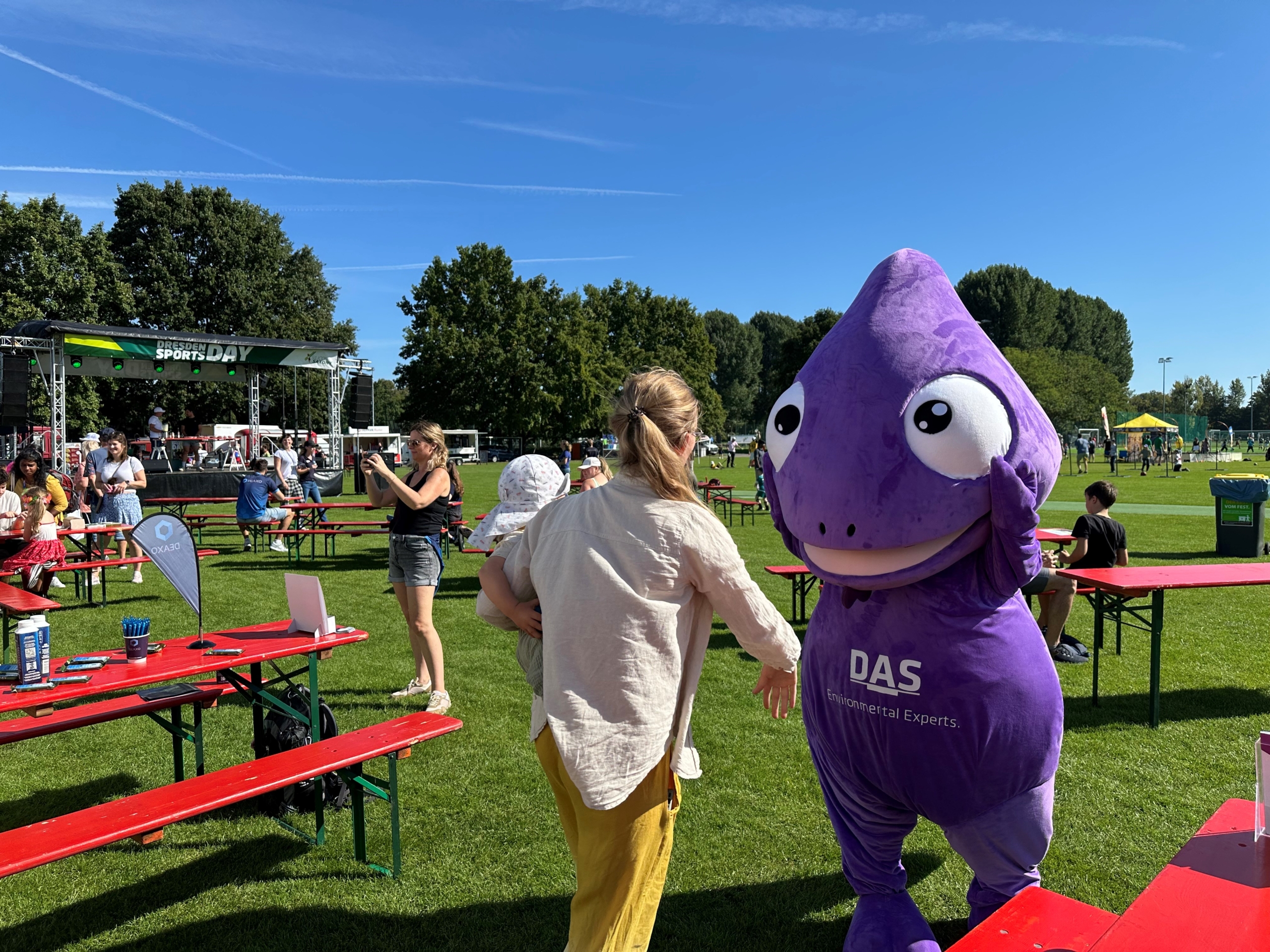 A person interacts with a large purple chameleon mascot in a lively outdoor event featuring green lawns, red picnic tables, and a stage under a clear blue sky, surrounded by attendees enjoying the sunny day.