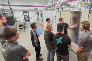 ISC Opening - 1 In einem technischen Schulungsraum stehen acht Personen um Schaltschränke, während eine Person Informationen erklärt; im Hintergrund sind komplexe Rohrleitungen und eine lila-weiße Wand sichtbar.