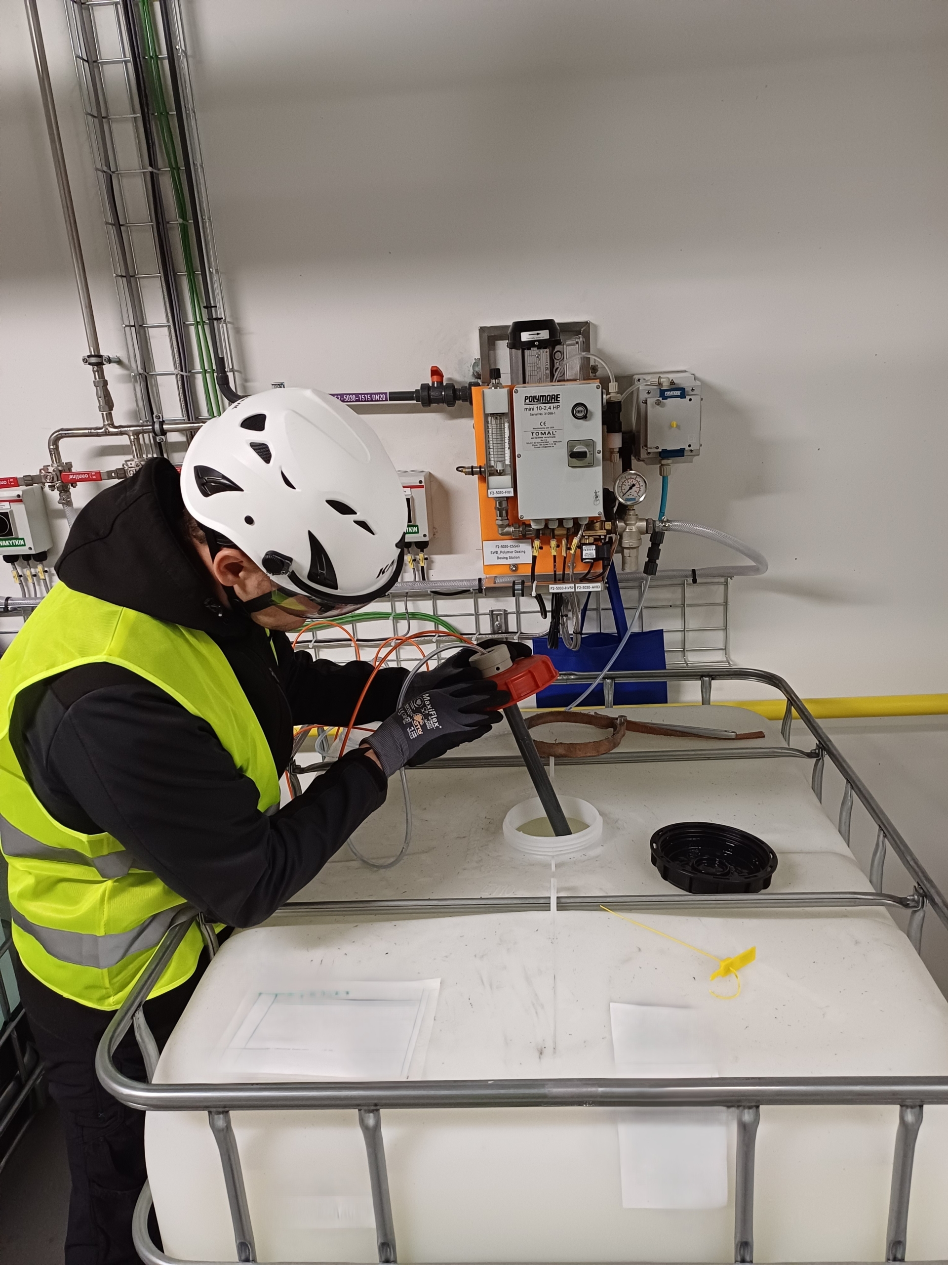 A worker in a safety vest and helmet examines a large liquid storage container with a control panel and pipes in the background, emphasizing industrial safety procedures.