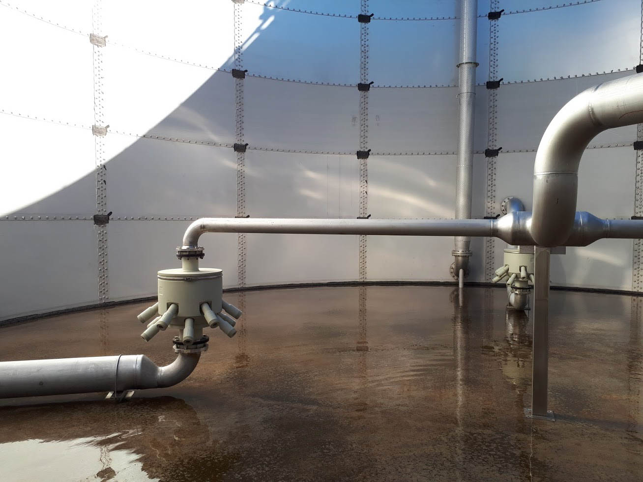 The interior of a large, empty industrial tank shows smooth metal walls and a floor with minimal water, featuring a network of shiny, silver pipes and valves prominently inside, related to maintenance or engineering systems.