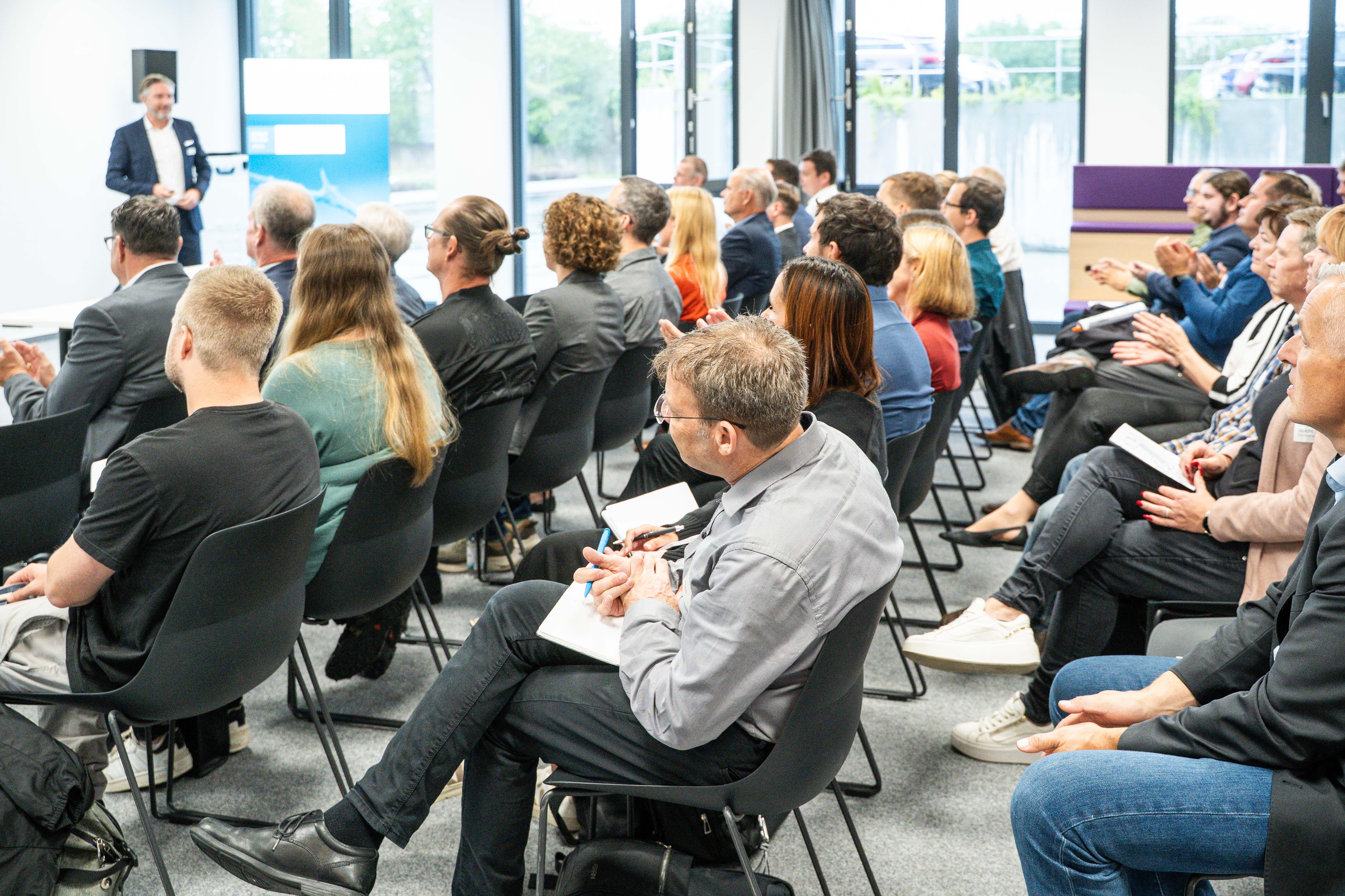 Audience attentively listening to a speaker at a business conference in a modern, well-lit room with large windows, featuring a mix of professionals seated in rows on black chairs, taking notes and engaging with the presentation.