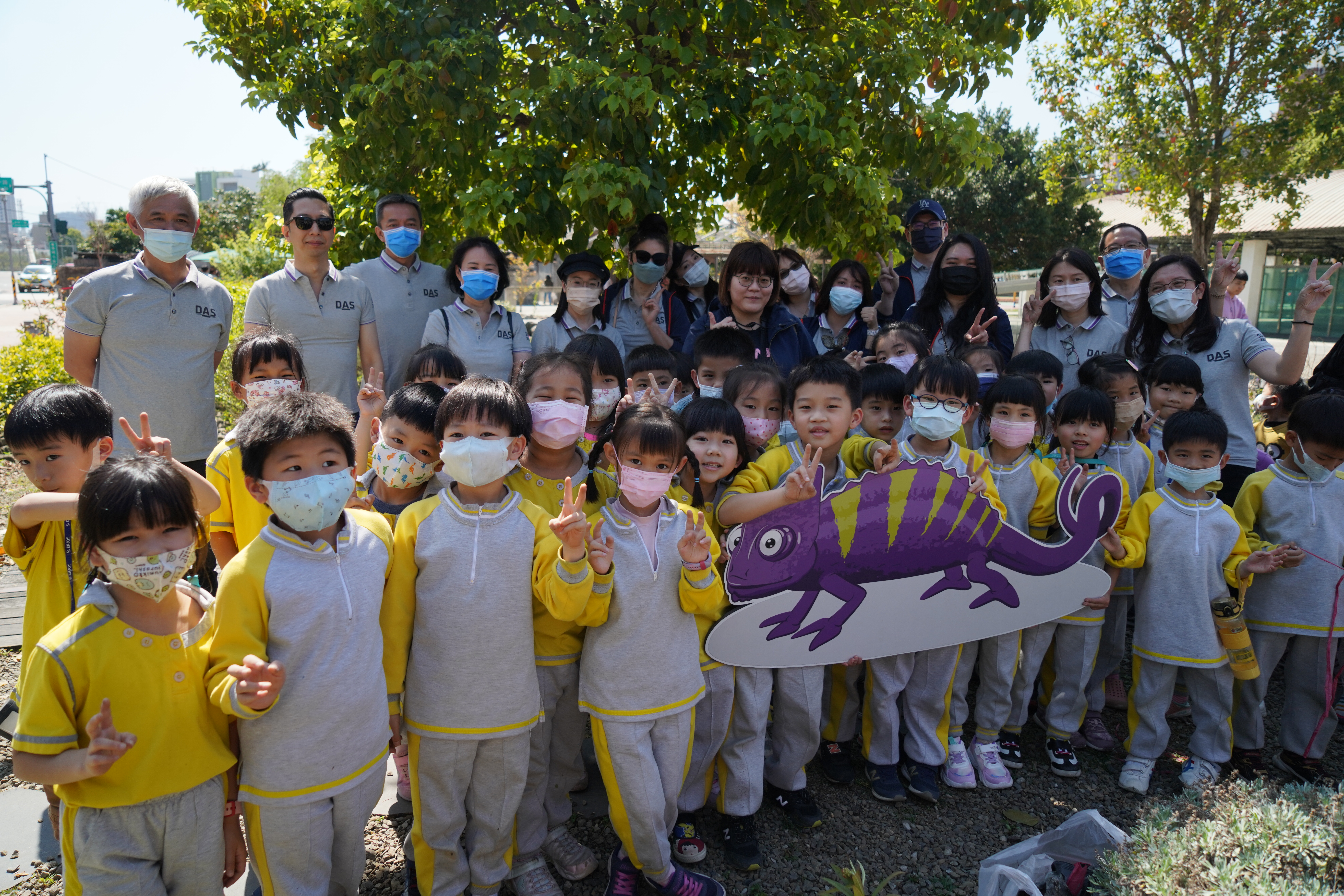 這張圖片顯示一群小學生在戶外活動中合影，孩子們穿著黃色和灰色的運動服，並戴著口罩，背景中有綠樹和成人陪伴者，顯示出快樂輕鬆的氛圍。