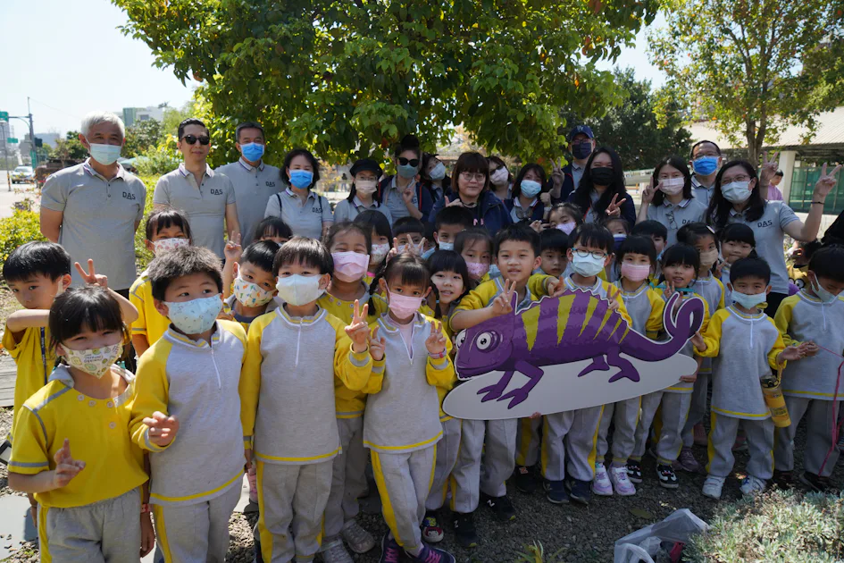 將軍村植樹活動 這張圖片顯示一群小學生在戶外活動中合影,孩子們穿著黃色和灰色的運動服,並戴著口罩,背景中有綠樹和成人陪伴者,顯示出快樂輕鬆的氛圍。
